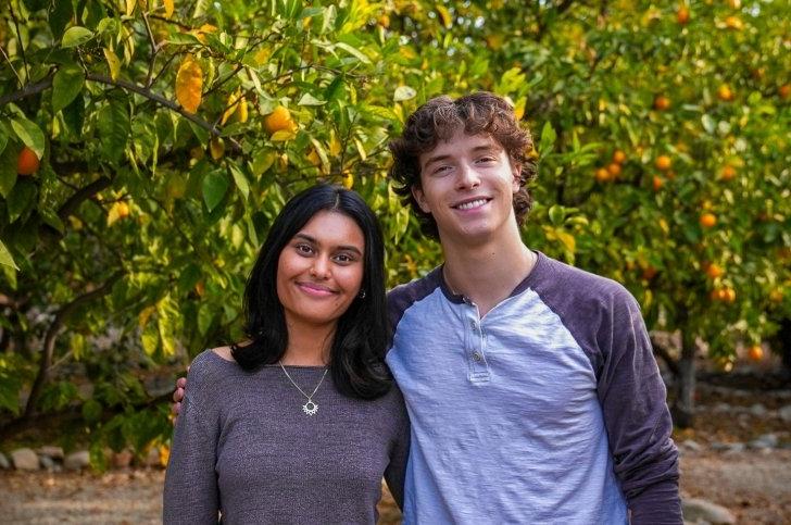 Sam McGinty and Aru Warrier smile and stand together in front of orange trees.